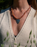 Collar Corazón puesto sobre un chaleco de lana crema: cordón anudado en naranjos y rojos con cuentas plateadas y un corazón con flor de lis al centro.