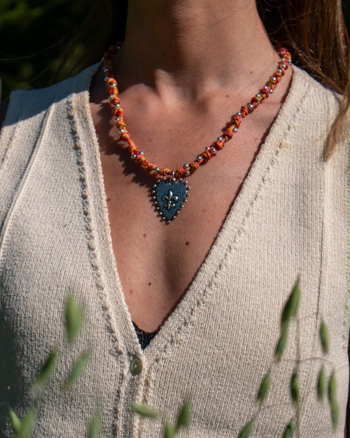 Collar Corazón puesto sobre un chaleco de lana crema: cordón anudado en naranjos y rojos con cuentas plateadas y un corazón con flor de lis al centro.